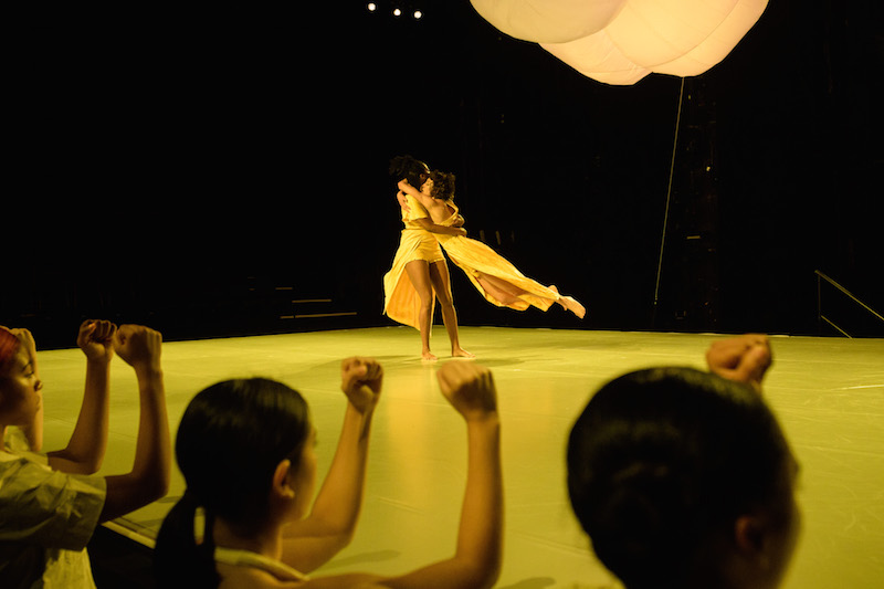 A dancer in marigold holds another artist and spins her around. A illuminated large lantern floats above them